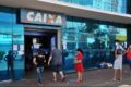 Agência da Caixa Econômica Federal com portas parcialmente abertas durante greve, clientes na fila e aviso de atendimento reduzido