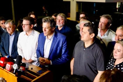 Renan Santos discursa em São Carlos com jovens ao fundo, cartazes sobre fim de isenções para unisquinas