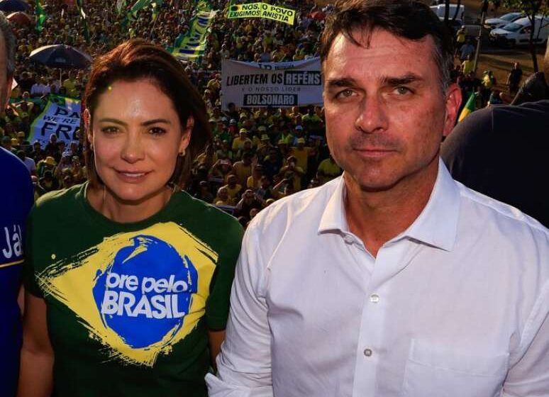 Flávio Bolsonaro discursa ao microfone com fundo desfocado, gesto de mão firme