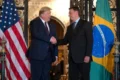 Ilustração política: Flávio Bolsonaro e Trump apertando as mãos com bandeira do Brasil subordinada à dos EUA