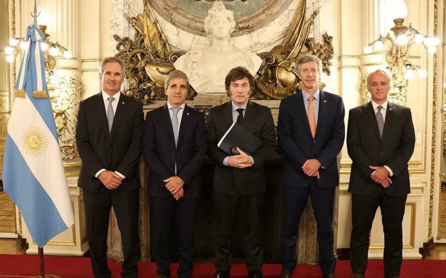 Javier Milei discursa com bandeira argentina ao fundo, gráficos de superávit fiscal e inflação em queda contrastam com silhuetas de aposentados e trabalhadores