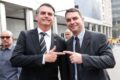Flávio Bolsonaro e Donald Trump apertando as mãos com bandeiras do Brasil e EUA ao fundo, simbolizando alinhamento político