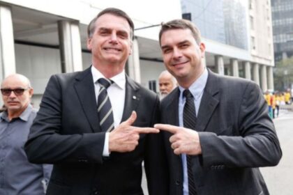 Flávio Bolsonaro e Donald Trump apertando as mãos com bandeiras do Brasil e EUA ao fundo, simbolizando alinhamento político