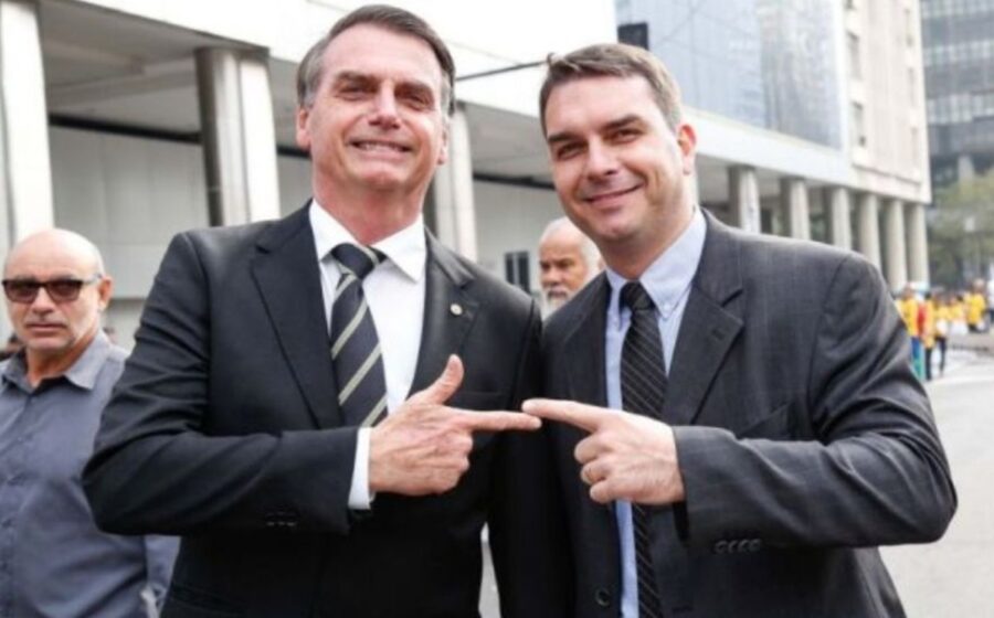 Flávio Bolsonaro e Donald Trump apertando as mãos com bandeiras do Brasil e EUA ao fundo, simbolizando alinhamento político