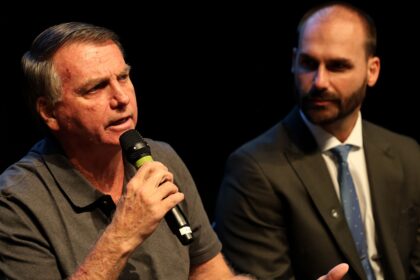 Senador Flávio Bolsonaro em sessão no Congresso Nacional, olhar sério, terno escuro, bandeira do Brasil ao fundo