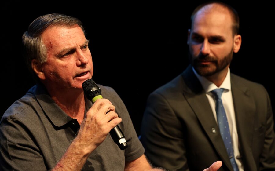 Senador Flávio Bolsonaro em sessão no Congresso Nacional, olhar sério, terno escuro, bandeira do Brasil ao fundo