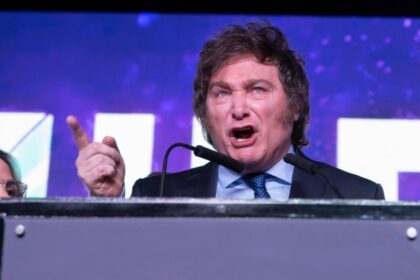 Javier Milei discursa enquanto gráficos mostram superávit fiscal e inflação em queda, contrastando com fila de argentinos em situação de vulnerabilidade