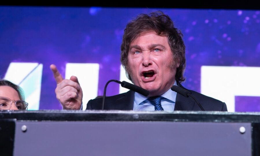 Javier Milei discursa enquanto gráficos mostram superávit fiscal e inflação em queda, contrastando com fila de argentinos em situação de vulnerabilidade