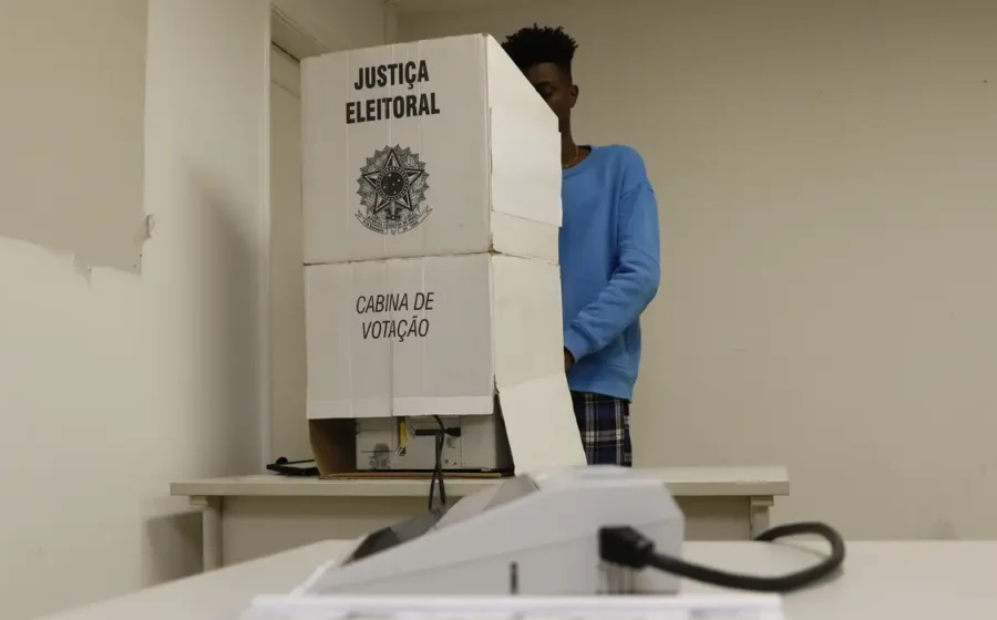 Eleitor na seção eleitoral com urna eletrônica e aviso "Proibida boca de urna" em destaque