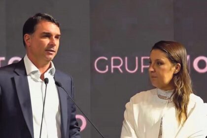 Flávio Bolsonaro discursa com logotipo da Caixa Econômica Federal e imagem de comunidade periférica ao fundo
