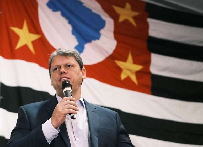 Governador com celular diante de obra paralisada e pedágio suspenso, simbolizando populismo que afugenta investimentos