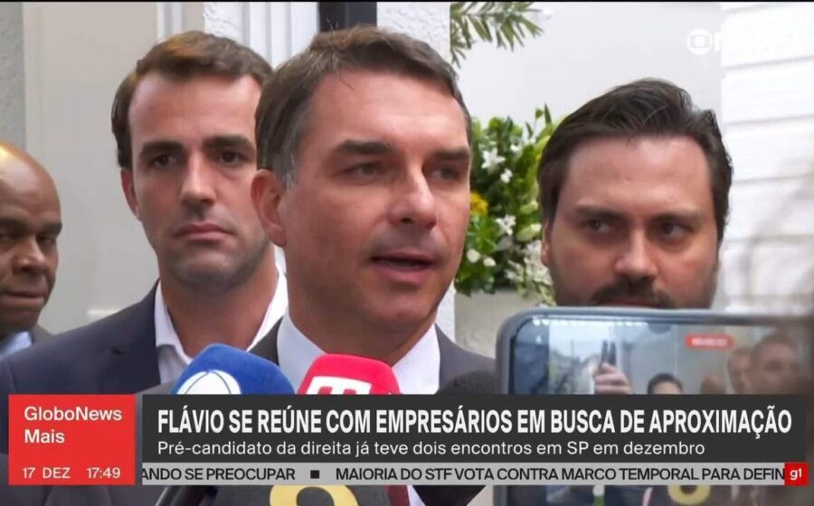 Flávio Bolsonaro em debate na Band com gráficos financeiros ao fundo