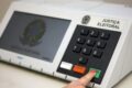 urna eletrônica sendo auditada por oficiais em mesa de votação com lacres e documentos