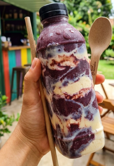 Garrafa PET transparente com açaí cremoso roxo, tampa rosa, na areia da praia de Guarapari